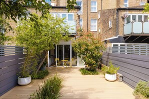 A courtyard with a wooden deck, potted plants, and a glass door entrance. - Colourful 3BDR Garden Maisonette in North London (Greater London)
