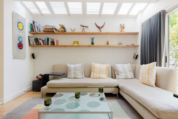 A modern living room with a sectional sofa, a glass coffee table, and a bookshelf with various items. - Colourful 3BDR Garden Maisonette in North London (Greater London)