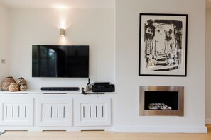 A living room with a flat-screen TV mounted above a white entertainment unit, a framed black and white artwork above a fireplace, and a small shelf with decorative items. - Colourful 3BDR Garden Maisonette in North London (Greater London)