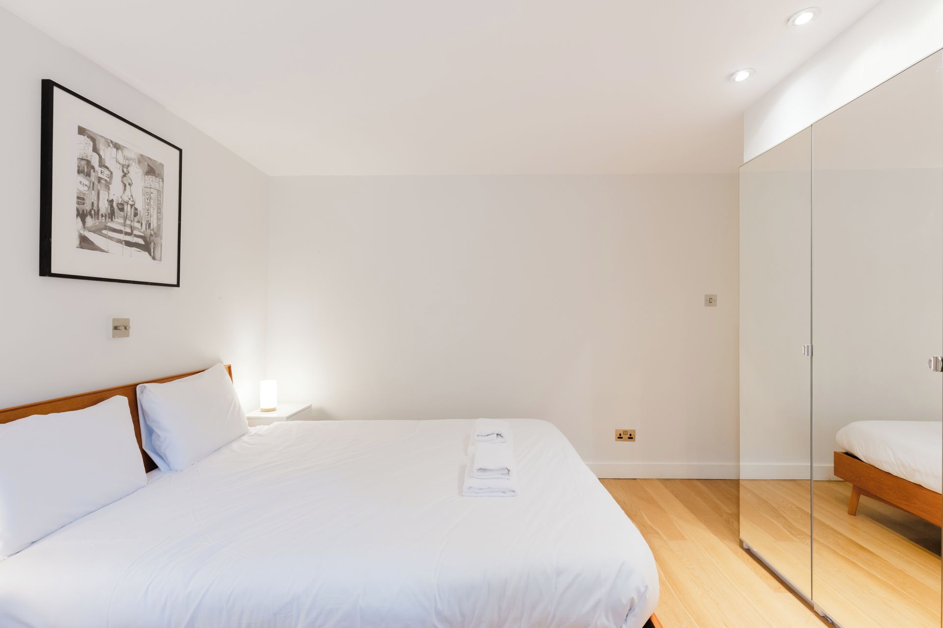 A modern bedroom with a large bed, a bedside table with a lamp, a framed picture on the wall, and a glass partition.