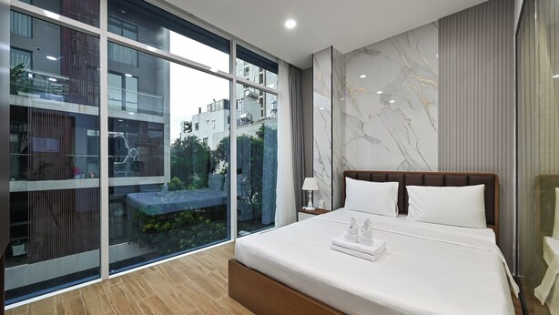 Una habitación de hotel moderna con una cama grande, una mesita de noche, una lámpara y vistas de la ciudad a través de ventanales del suelo al techo.