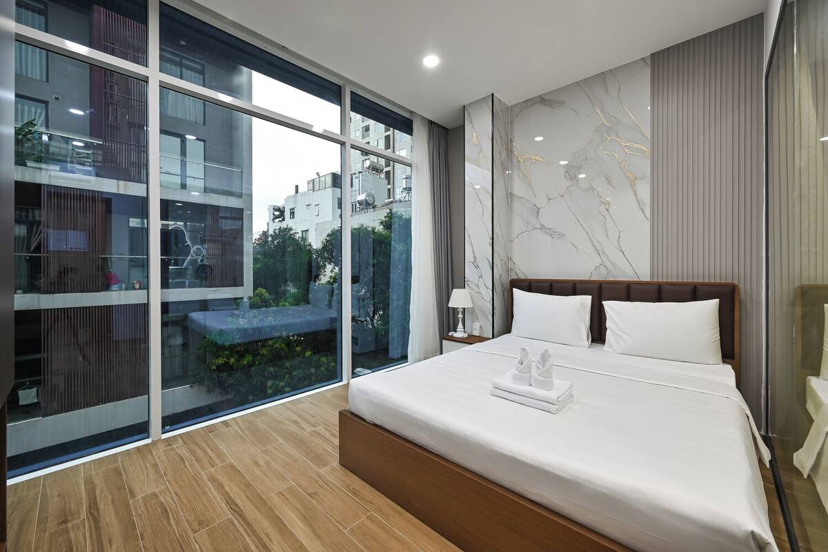 A modern hotel room with a large bed, a nightstand, a lamp, and a view of the city through floor-to-ceiling windows.