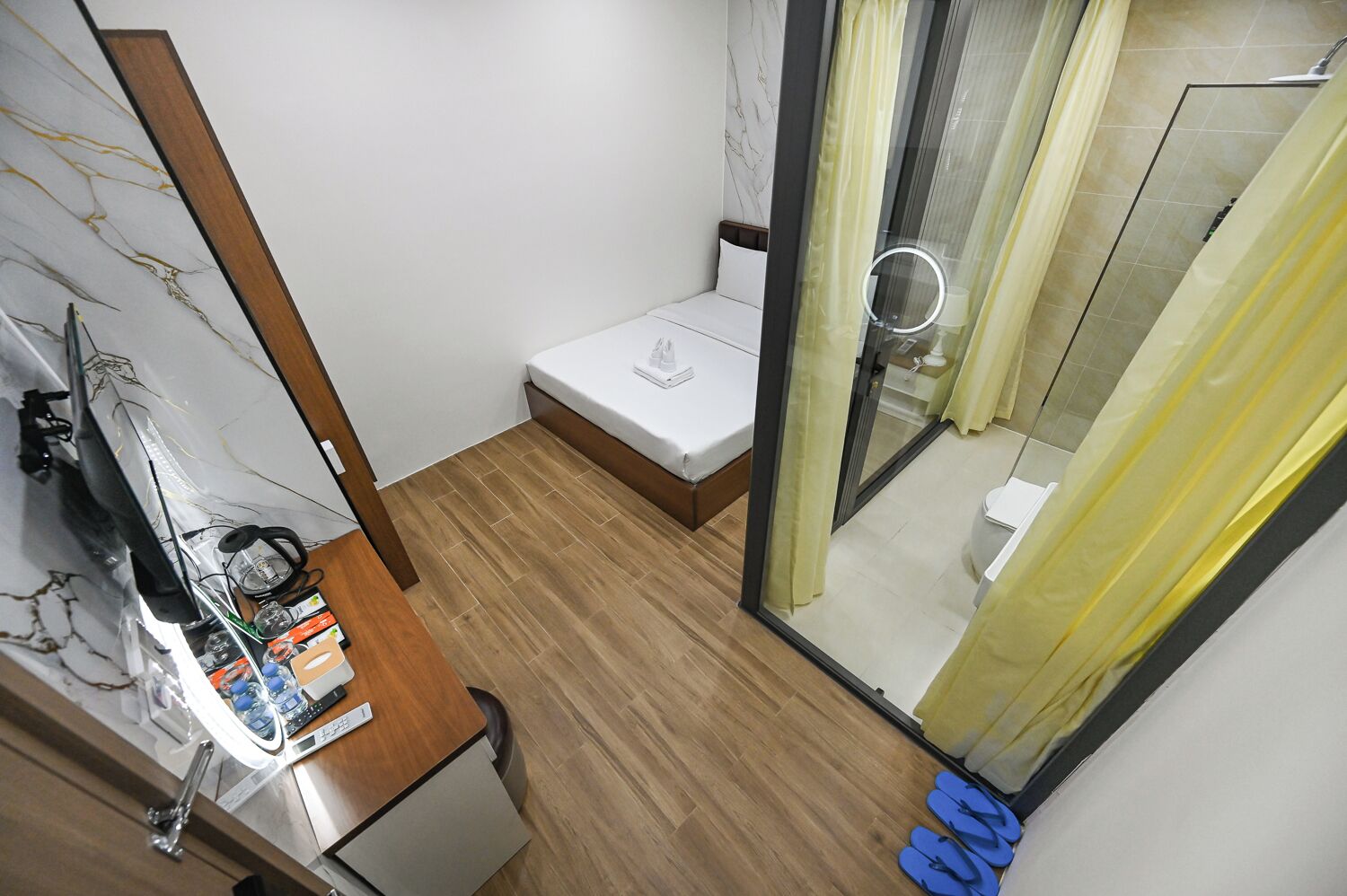 A compact hotel room with a wooden floor, a bed, a small bedside table with a lamp, and a shower area with a glass door and yellow curtains.