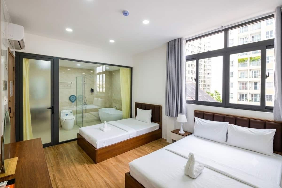 A hotel room with a large bed, a smaller bed, a glass shower enclosure, and a view of buildings through the window.