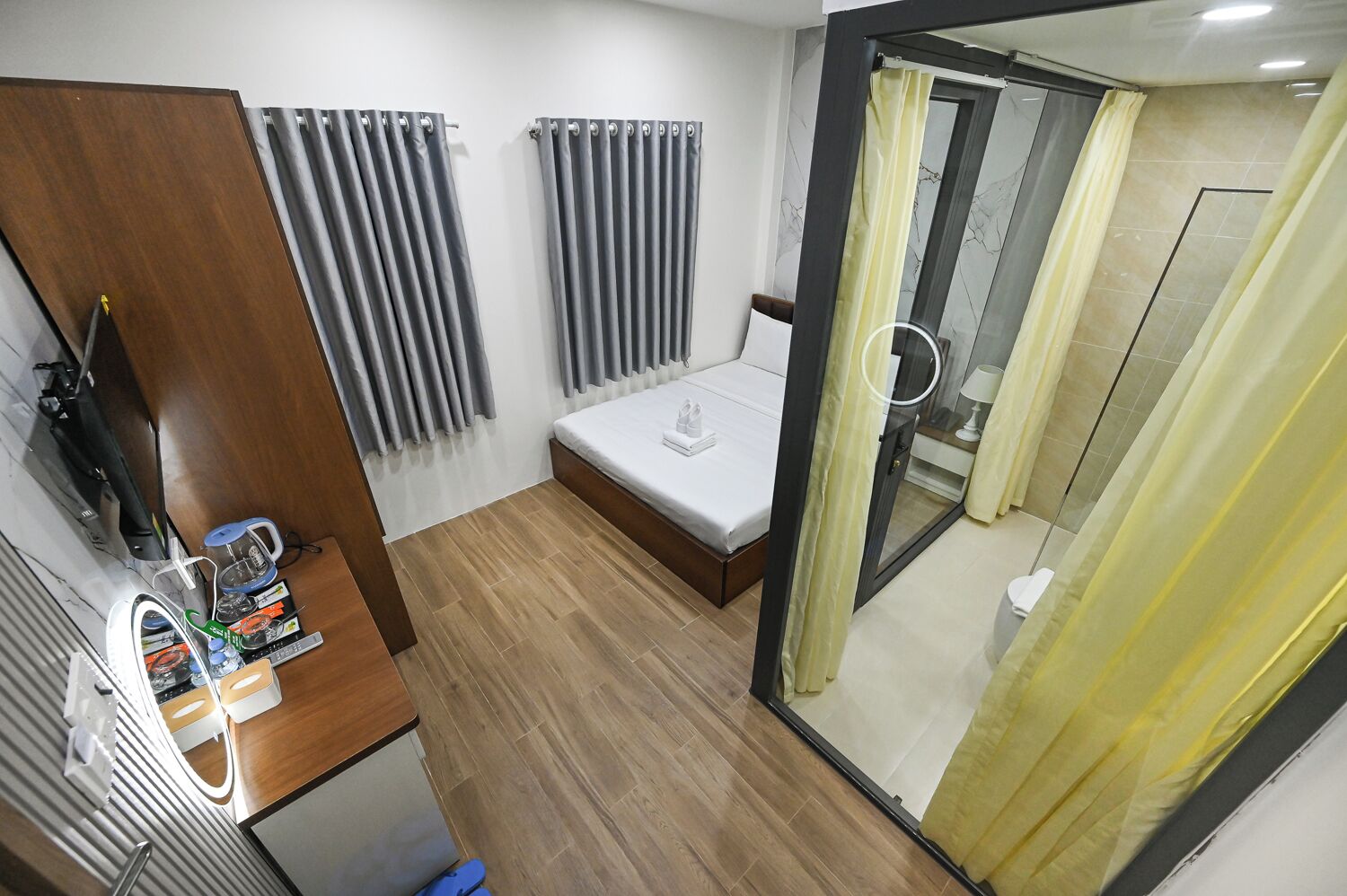 A compact hotel room with a bed, a small desk with a chair, a shower area, and a view through a glass door.
