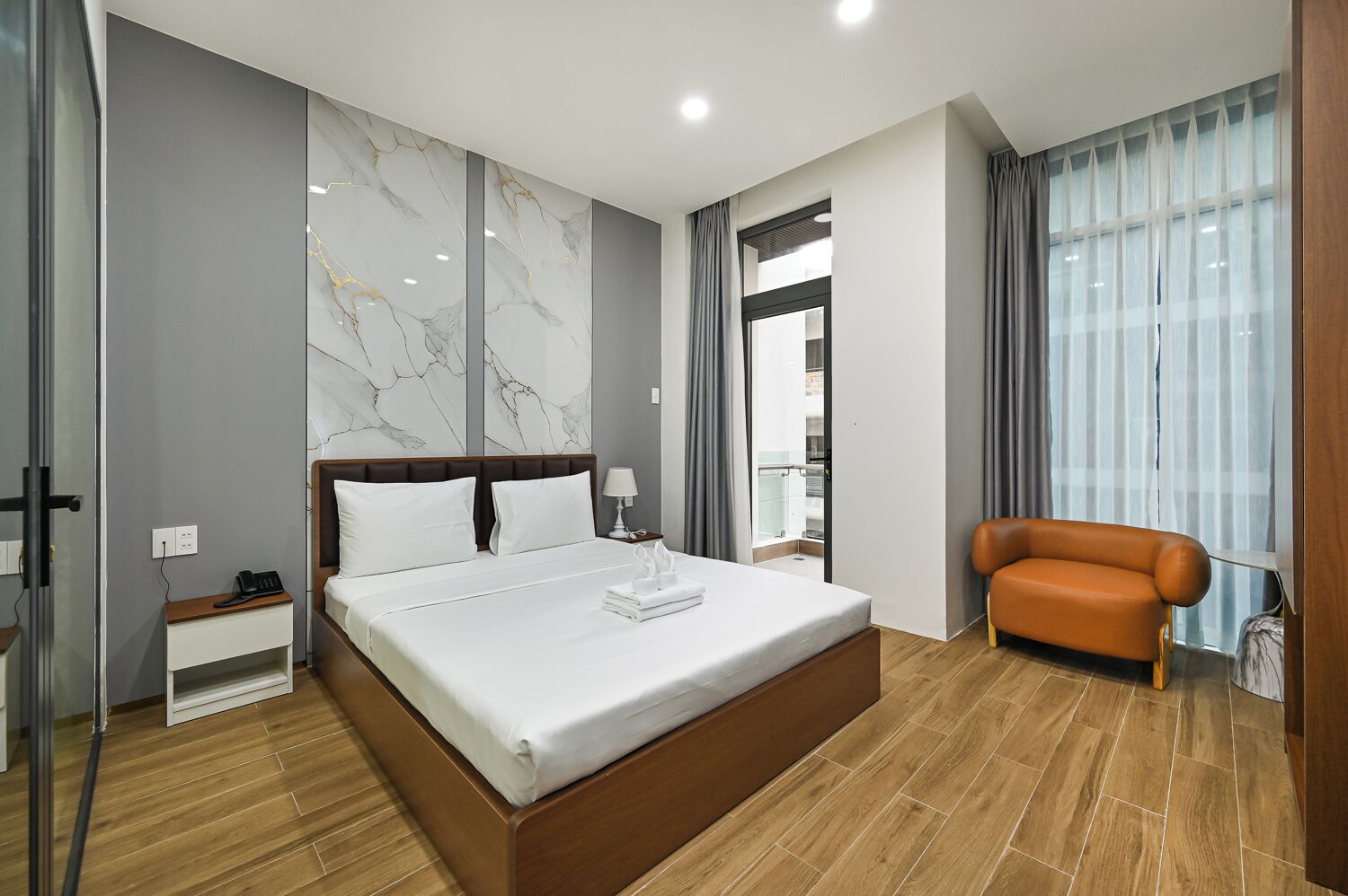 A modern hotel room with a large bed, a wooden nightstand, a chair, and a view of the city through the window.