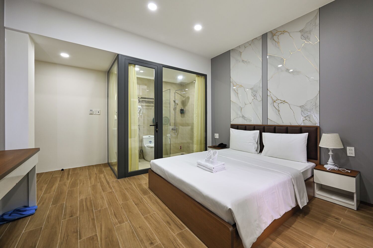 A hotel room with a bed, bedside table, nightstand, and a glass-enclosed shower area.