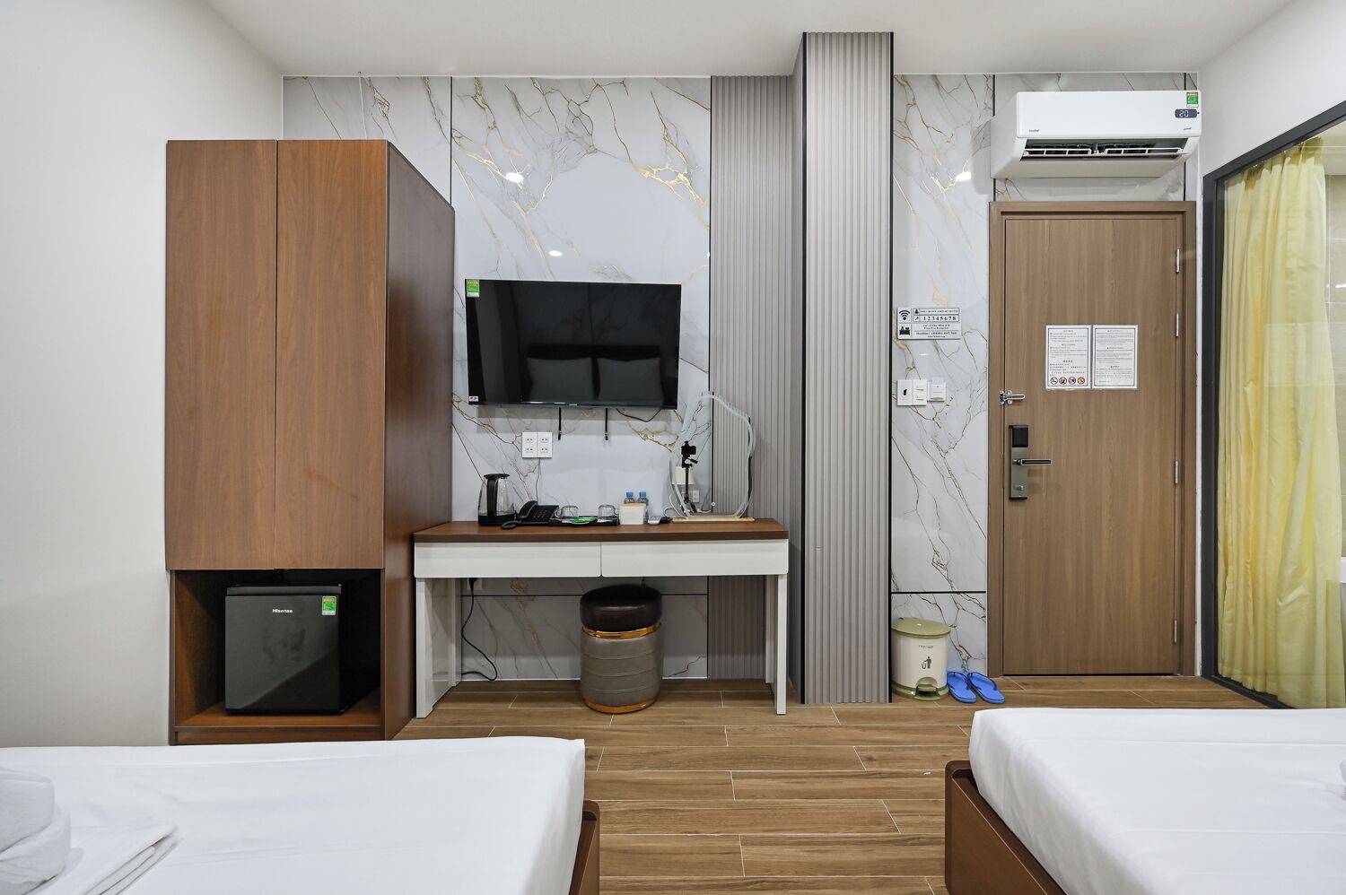 A hotel room with a bed, a desk with a TV, a small fridge, a trash bin, and a door with a sign.