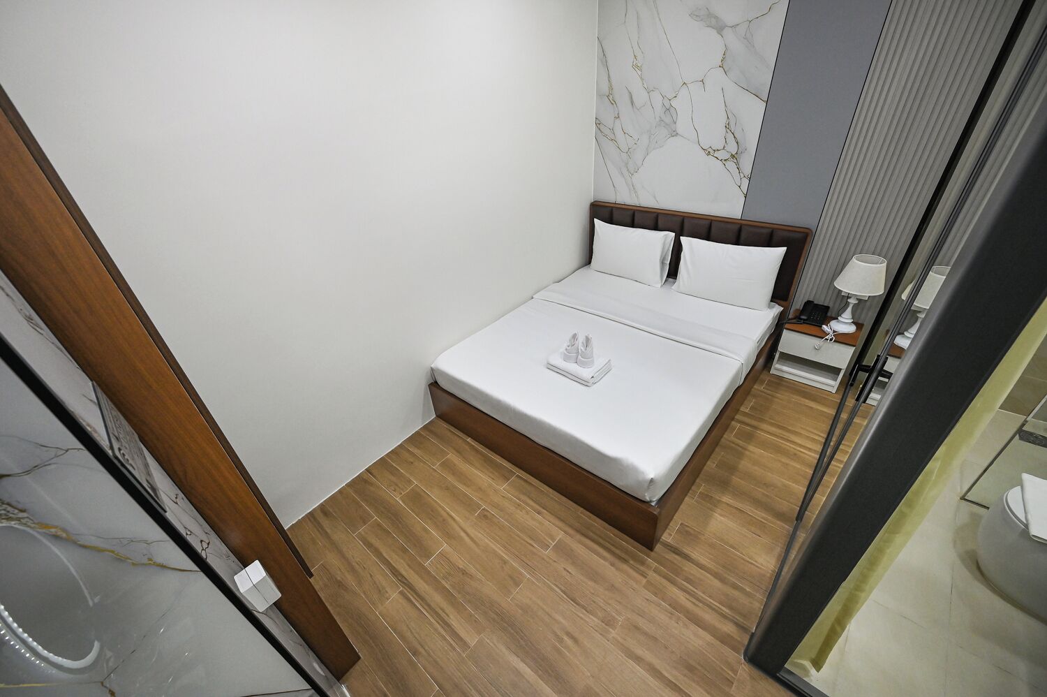 A hotel room with a bed, a marble wall, a wooden floor, and a bedside table with a lamp.