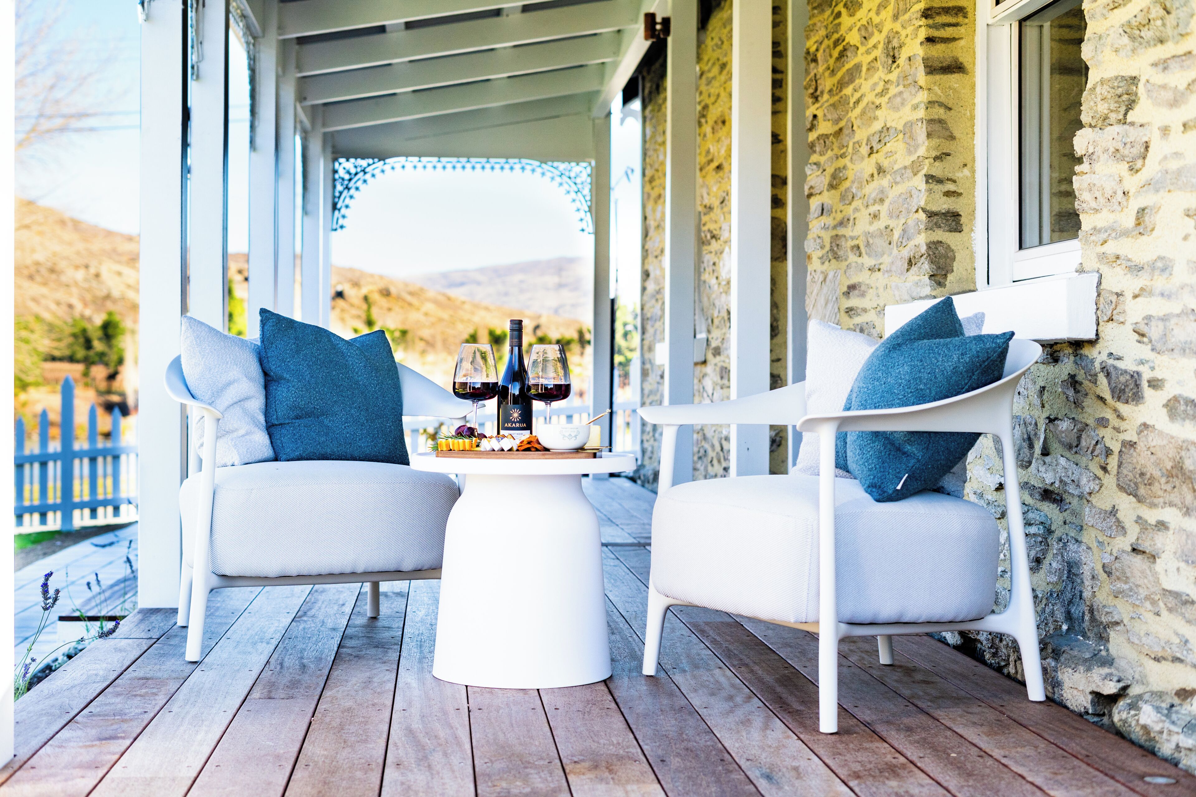 A covered outdoor seating area with two white chairs and a table, set against a stone wall with a view of the hills.