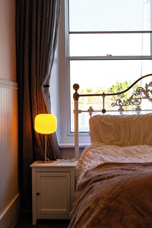 A bedroom with a bed, a nightstand with a lamp, a window with curtains, and a view of greenery outside.
