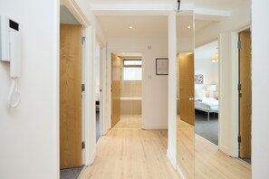 Een gang met houten vloer, een badkamer met bad en een slaapkamer met bed en nachtkastje.