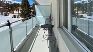 Un balcón con barandilla de vidrio, una mesa y una silla. Se ve una vista de montañas nevadas y casas al fondo.