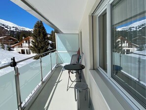 Ein Balkon mit Glasgeländer, einem Tisch und einem Stuhl. Im Hintergrund sind schneebedeckte Berge und Häuser zu sehen.