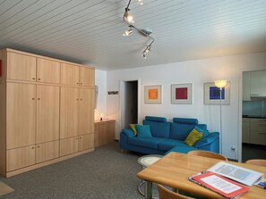 Ein gemütlicher Wohnraum mit einem Holzschrank, einem blauen Sofa, einem Esstisch mit einem Buch und einer Deckenleuchte.