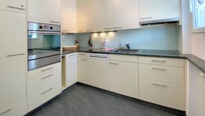 Una cocina moderna con armarios blancos, horno de acero inoxidable, lavavajillas y una ventana con cortinas traslúcidas.