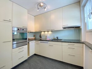 Eine moderne Küche mit weißen Schränken, einem Edelstahlbackofen, einem Geschirrspüler und einem Fenster mit durchsichtigen Vorhängen.