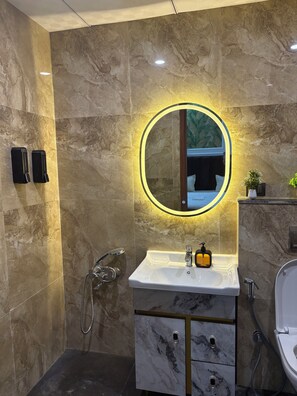 Une salle de bain moderne avec un mur en marbre, un grand miroir rond avec éclairage intégré, un lavabo blanc, une douchette à main et des toilettes.