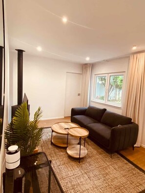 Ein modernes Wohnzimmer mit einem dunklen Sofa, zwei runden Holztischen, einem Glastisch und einer Zimmerpflanze, sowie einem großen Fenster mit Vorhängen.