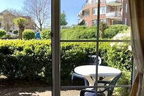 Blick aus einem Fenster auf eine Wohngegend mit einer Hecke, einem weißen Tisch und Stühlen sowie einem blauen Auto im Hintergrund.