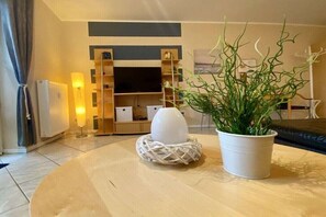 Ein Wohnzimmer mit rundem Tisch, einer Topfpflanze, einem gewebten Korb, einer weißen Vase und einer TV-Konsole mit Regalen.