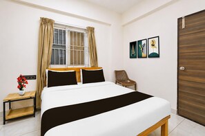 A hotel room with a bed, a window with curtains, a chair, a small table with a vase, and artwork on the wall.