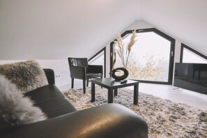 A modern living room with a black leather sofa, a black armchair, a small table, and a large window with a view of trees. - Bildstein's "Kathmandu" (Langenfeld, Deutschland)