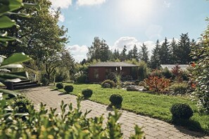 A red building with a sloped roof, surrounded by a well-maintained garden with various shrubs and a paved pathway leading to the entrance. - Bildstein's "Kathmandu" (Langenfeld, Deutschland)