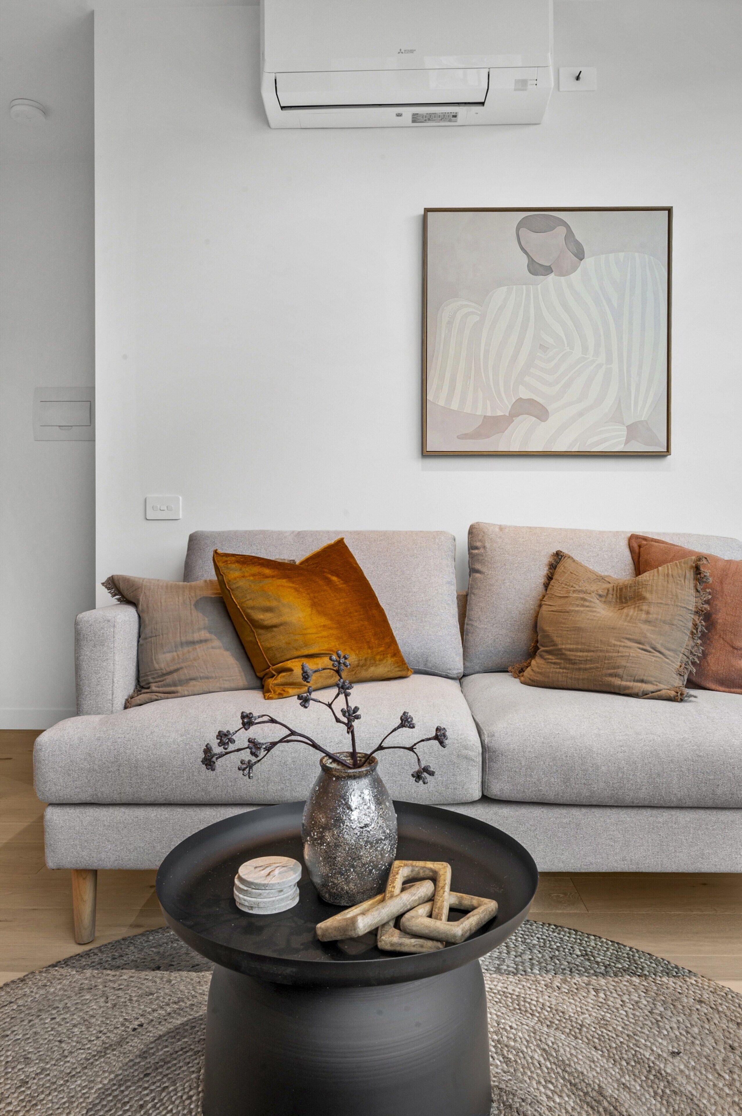 Een moderne woonkamer met een grijze bank, decoratieve kussens, een zwarte salontafel met een vaas en houten objecten, en een lijst met een abstract schilderij aan de muur.