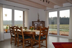 A dining room with a table set for six, wooden chairs with green cushions, a wooden cabinet with a vase of flowers, and large windows with star decorations. - Holiday apartment with 4 bedrooms (Greven)