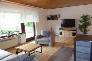 Un salon avec un canapé, une table basse, un téléviseur, une plante et une fenêtre avec des rideaux.