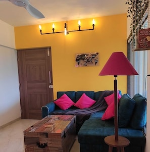 A living room with a blue sofa, a wooden coffee table, a red floor lamp, a world map wall art, and a ceiling fan. - Chillout space-Palghar with private JACUZZI! (Velgaon)