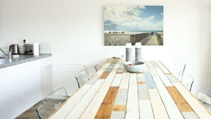 Et spiseområde med et langt træbord, stole, en hvid radiator, en køkkenbordplade med hårde hvidevarer, og et maleri af en strand på væggen.