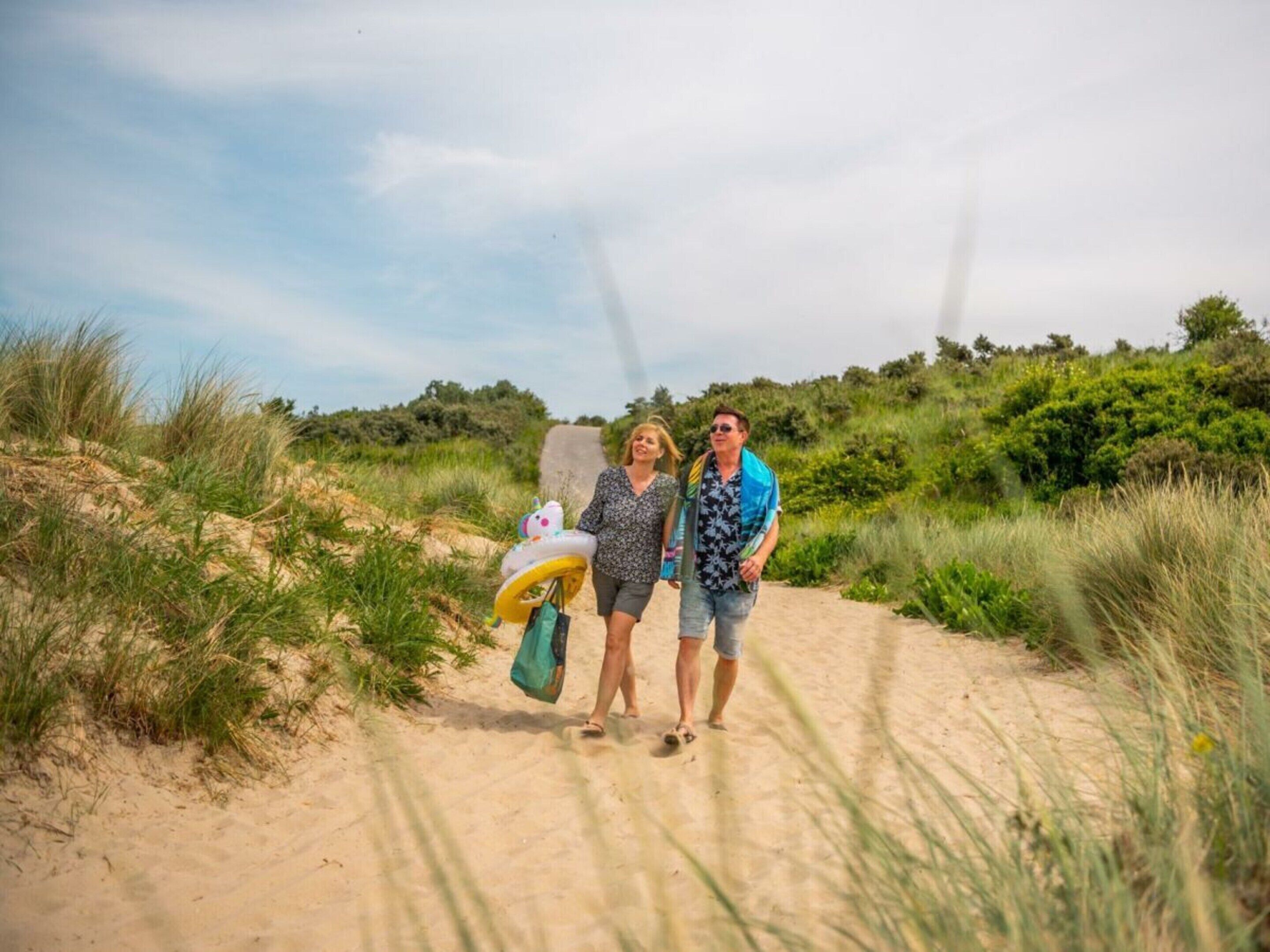 Uma trilha de areia com vegetação ao longo dos dois lados, uma pessoa carregando uma sacola e outra com uma prancha de surf.