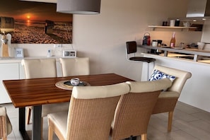 Ein Essbereich mit Holztisch und Korbstühlen, eine Küche mit modernen Geräten und ein Wandbild, das einen Sonnenuntergang über einem Strand zeigt.