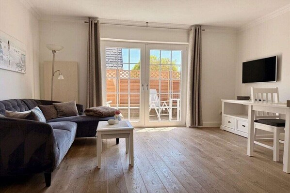 En stue med sofa, et lille bord, et fjernsyn og en skydedør, der fører til et udendørs område med et træhegn.