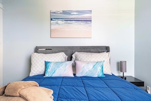A bedroom with a bed, pillows, a blanket, a nightstand, a lamp, and a beach view painting. - Jax Beach Zen Retreat – Walk to Ocean & Pier (Jacksonville Beach)