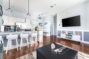 A modern kitchen with white cabinets, a large island with bar stools, a flat-screen TV mounted on the wall, and a dining area with a black table. - Jax Beach Zen Retreat – Walk to Ocean & Pier (Jacksonville Beach)