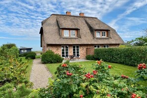 Ein Haus mit einem Reetdach, verbunden mit einem gepflasterten Weg, umgeben von üppigem Grün und blühenden Pflanzen.