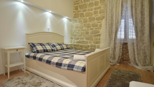 A bedroom with a stone wall, a bed with a plaid bedspread, a nightstand, a chandelier, and a window with curtains. - Studios Villa Rossella 4 (Rovinj)