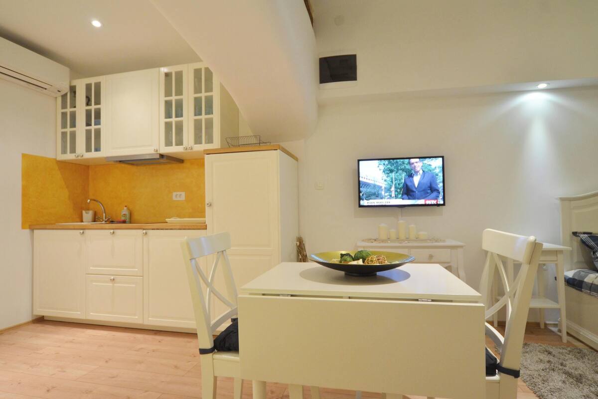 Uma cozinha compacta com armários brancos, uma mesa de jantar com cadeiras e uma televisão fixada na parede.