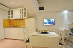 Uma cozinha compacta com armários brancos, uma mesa de jantar com cadeiras e uma televisão fixada na parede.