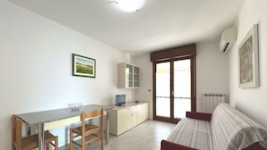 A compact living space with a kitchenette, a dining area, a sofa, and a large window providing natural light. - Well Equipped Holiday Home in Grado (Grado)