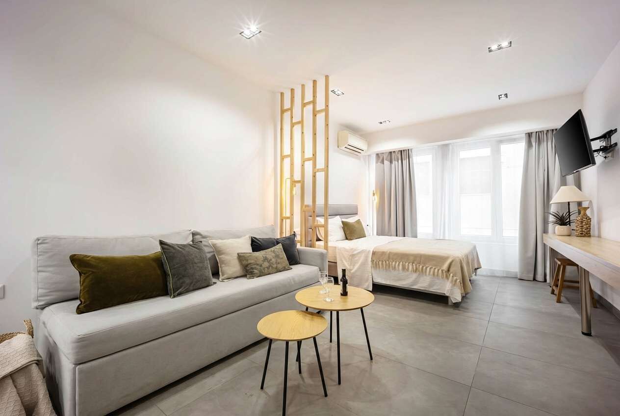 A modern hotel room with a sofa, a bed, a small round table, a TV mounted on the wall, and a window with curtains.