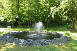 Ein runder Teich mit einer zentralen Fontäne, umgeben von einem Steinrand, in einem üppigen Grünpark.