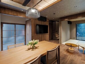Ein modernes Interieur mit einem Holzes Esstisch, einem an der Wand befestigten Fernseher und Blick ins Freie durch ein großes Fenster.