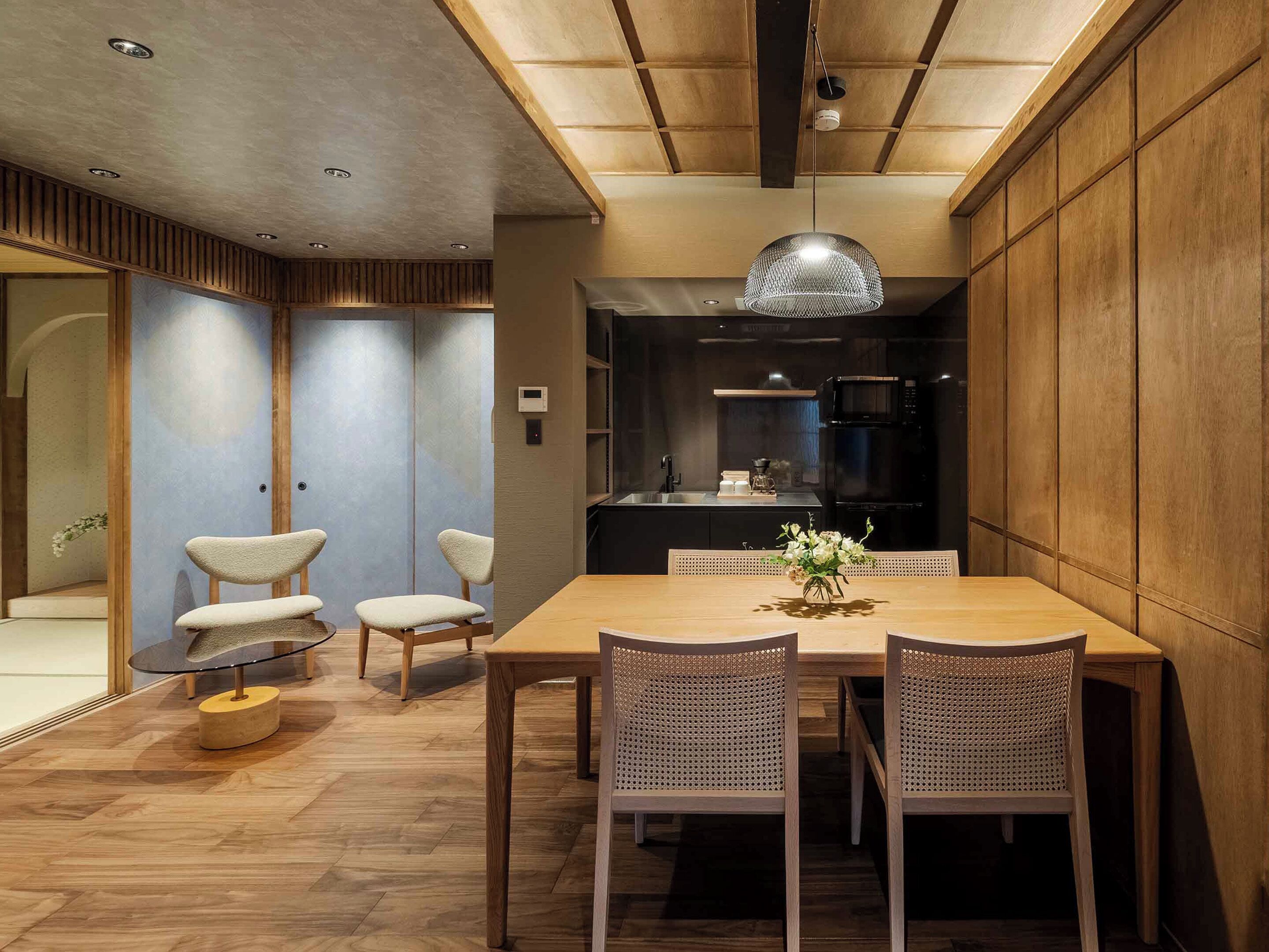 A modern dining area with a wooden table, four chairs, a hanging light fixture, and a kitchen in the background.