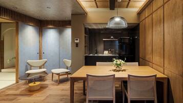 A modern dining area with a wooden table, four chairs, a hanging light fixture, and a kitchen in the background.