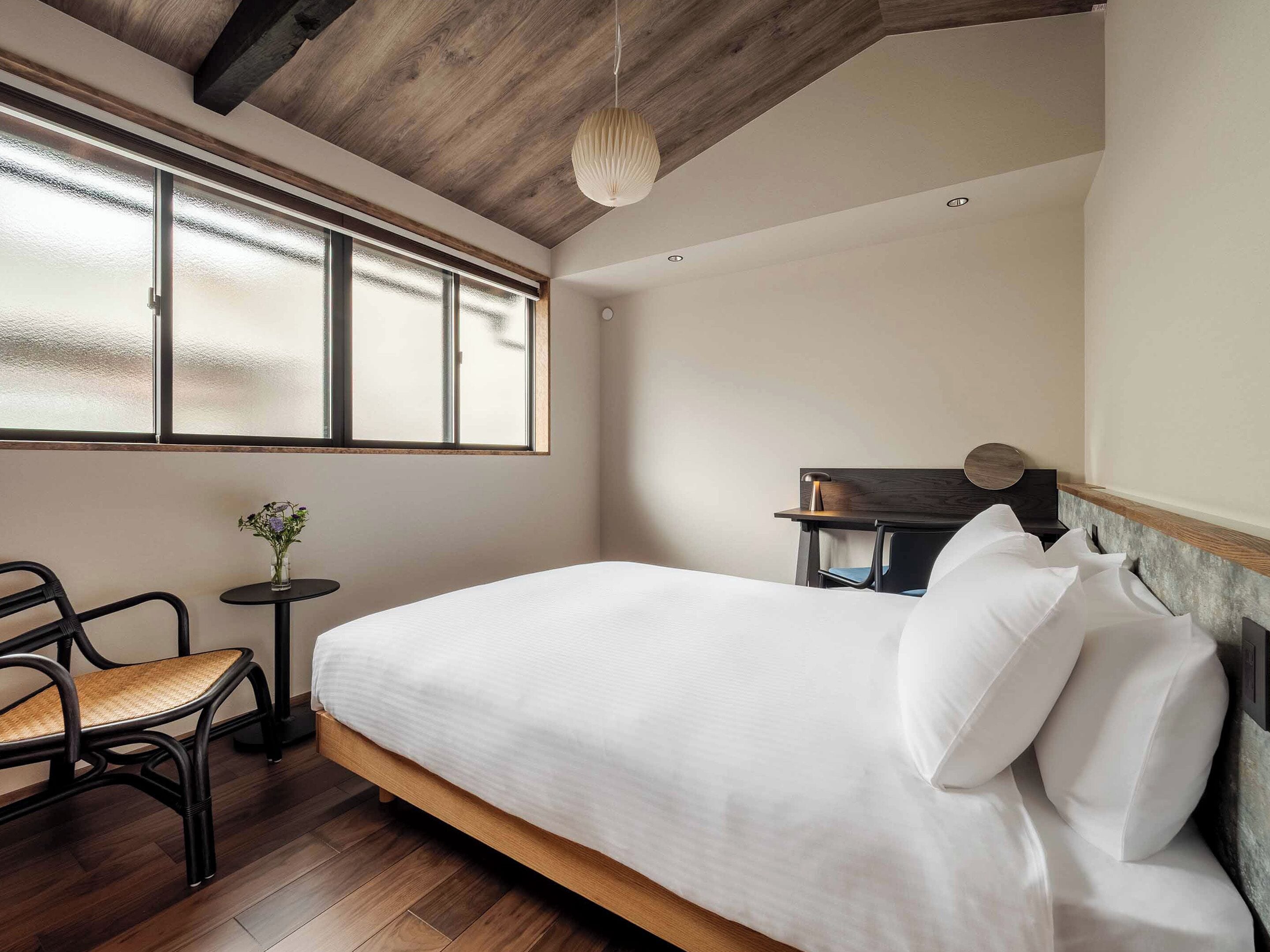 A bedroom with a bed, a chair, a small table with a vase, and a window with frosted glass.