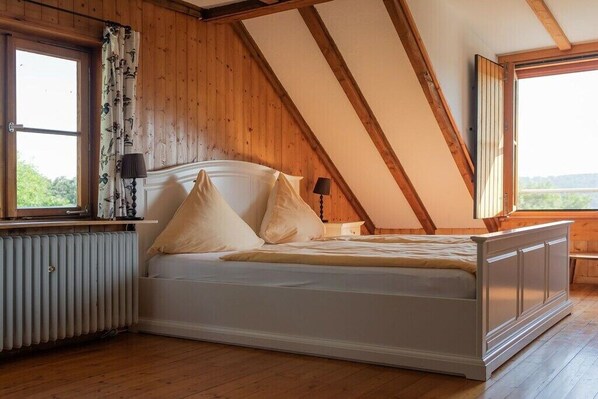 A bedroom with a sloped ceiling, a bed with pillows, a bedside lamp, and a window with curtains. - Gommerscheid Hunting Lodge (Ehlscheid)